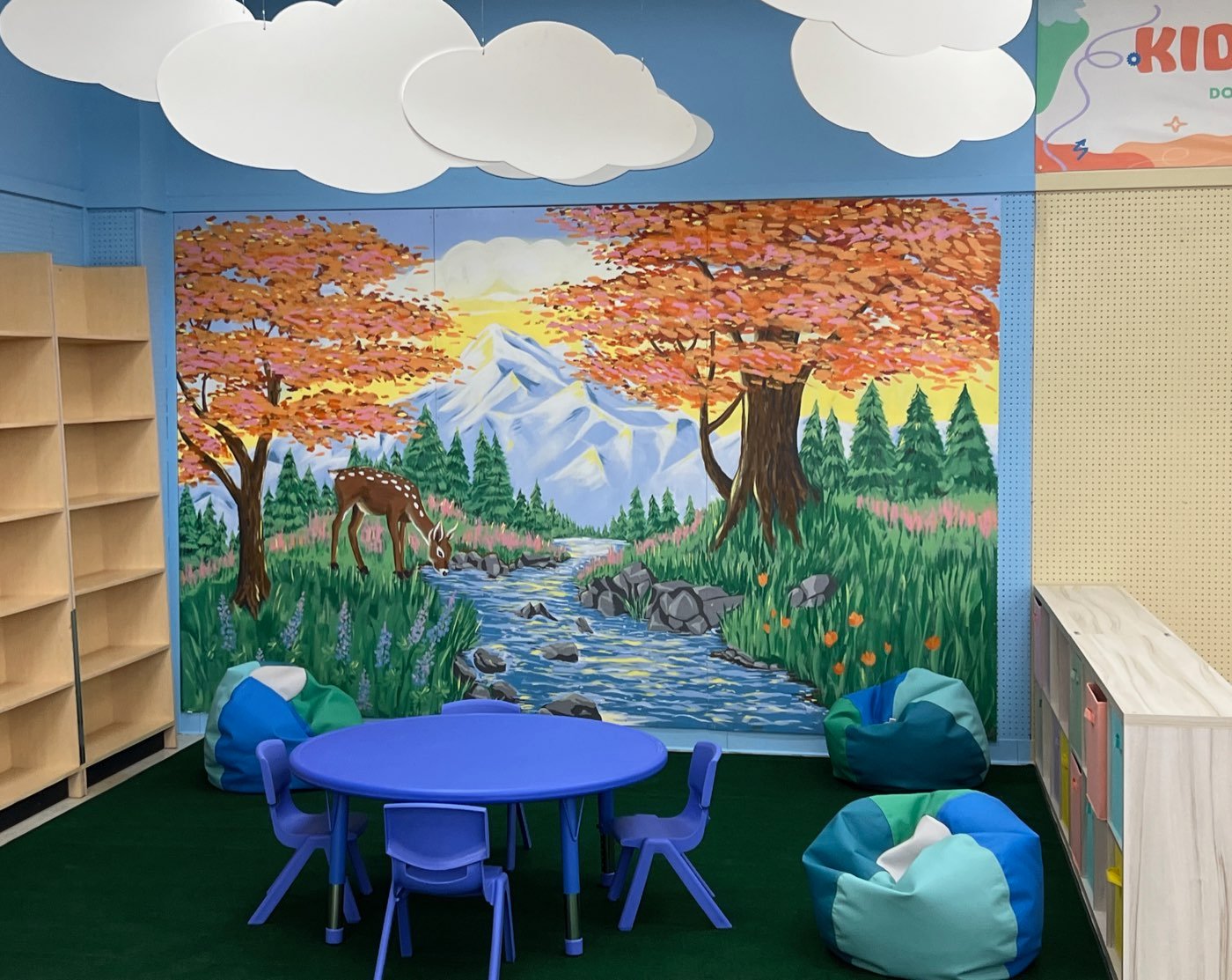 Painted mural of a deer drinking from a mountain stream beneath autumn trees, in a children's playspace with bean bags and a small table
